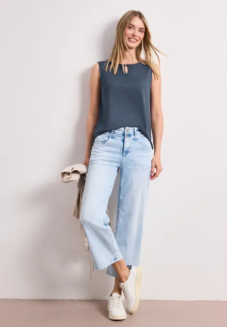 Eine Frau trägt ein ärmelloses dunkelblaues Oberteil, helle Jeans und weiße Sneakers vor hellem Hintergrund.