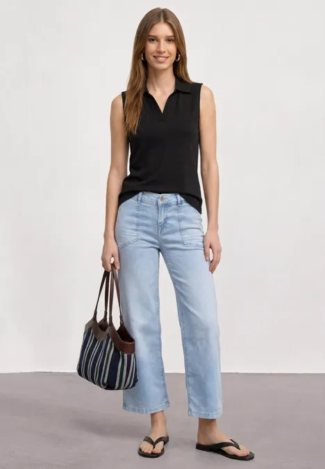 Frau in schwarzem Top, hellen Jeans, Flip-Flops, gestreifte Tasche, vor neutralem Hintergrund.