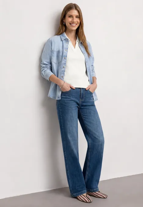 Top in Rippstruktur mit Polokragen off white Eine Frau lehnt an einer Wand, trägt helle Bluse, Jeanshemd, weite Jeans und gestreifte Schuhe.