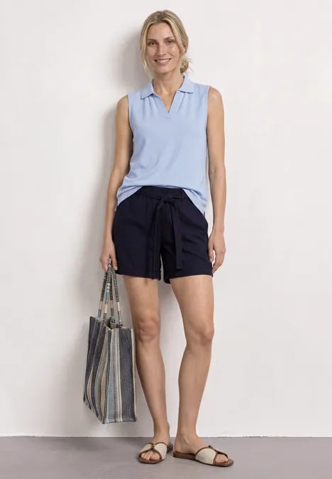 Vrouw in mouwloze lichtblauwe polo, donkerblauwe shorts, gestreepte tas en sandalen voor witte achtergrond.