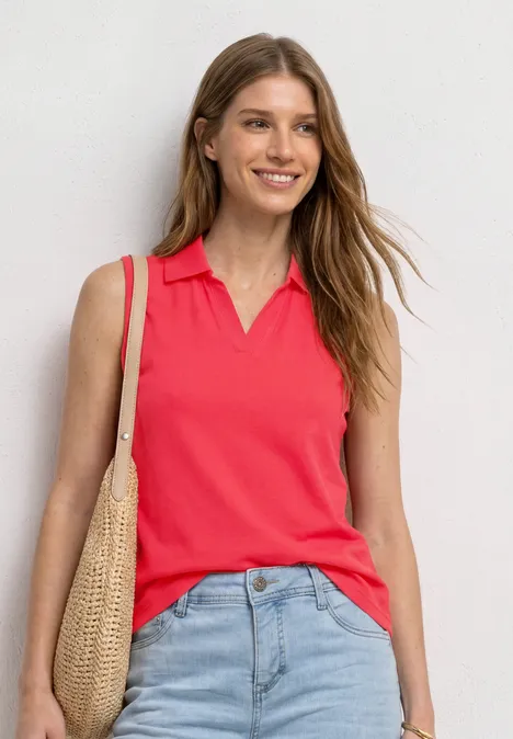 Frau vor weißer Wand in rotem ärmellosem Poloshirt, blauen Jeans und Korbtasche.