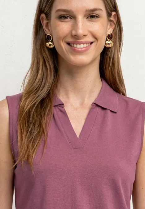 Eine Frau mit langen braunen Haaren trägt ein ärmelloses lila Poloshirt und goldene Creolen.