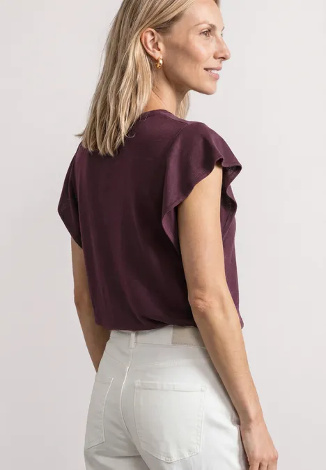 Shirt mit kurzen Volant-Ärmeln rustic rouge Frau in weißer Hose und lila Oberteil mit Rüschenärmeln, von hinten fotografiert