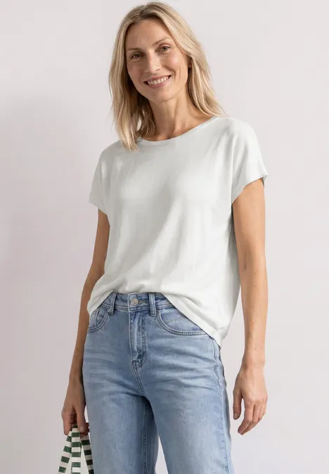 Femme en t-shirt blanc et jean bleu clair devant un fond neutre.