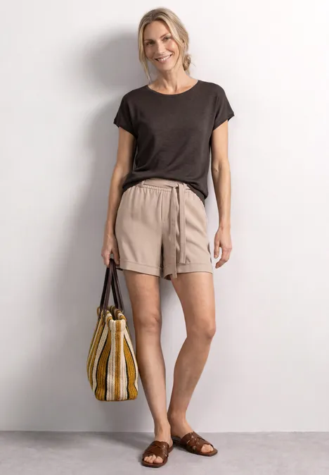 Chemise dropped shoulder à col rond espresso brown Femme en t-shirt noir, short beige et sandales marron, portant un sac rayé jaune et blanc.