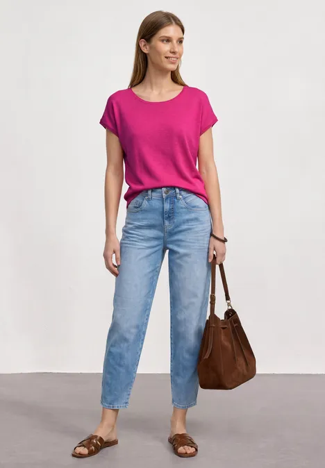 Frau in pinkem T-Shirt, blauen Jeans, brauner Tasche und Sandalen vor neutralem Hintergrund.