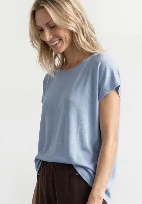 Dropped Shoulder Shirt mit Rundhals cashmere blue Frau mit blondem Haar trägt lässiges hellblaues T-Shirt und dunkle Hose vor hellem Hintergrund.