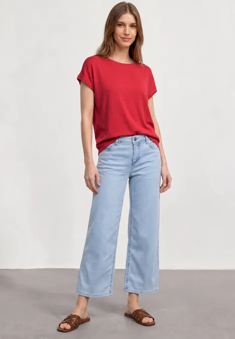 Frau trägt rotes T-Shirt, hellblaue weite Jeans und braune Sandalen vor neutralem Hintergrund