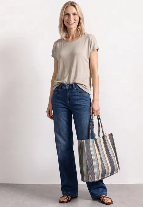 Frau im beigen T-Shirt, blauen Jeans und schwarzen Schuhen mit gestreifter Einkaufstasche.