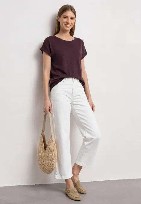 Dropped Shoulder Shirt mit Rundhals rustic rouge Frau in dunkelviolettem T-Shirt, weißen 7/8-Hosen und beigen Slippern mit Strohtasche, vor weißer Wand.