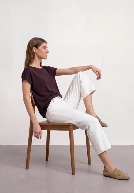 Dropped Shoulder Shirt mit Rundhals rustic rouge Junge Frau sitzt auf einem Stuhl, trägt ein bordeauxfarbenes T-Shirt, weiße Hose und beige Slipper.
