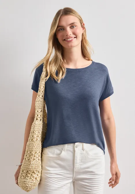 Frau in blauem T-Shirt und weißer Hose mit Basttasche über der Schulter.