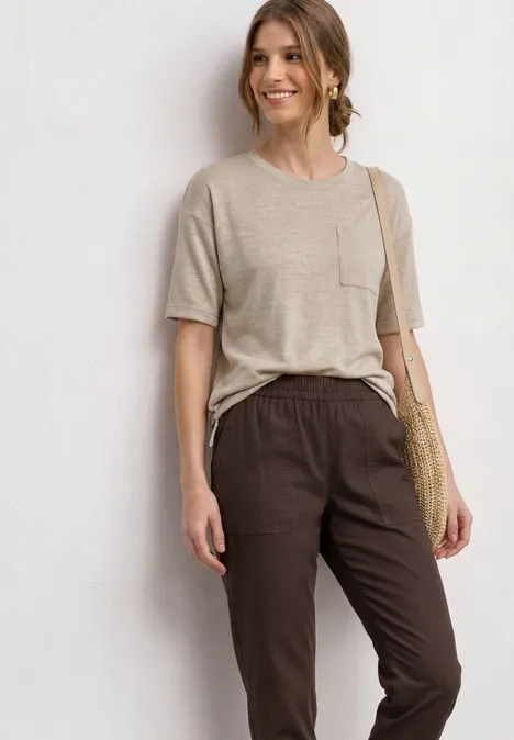 T-Shirt mit Rundhals und Brusttasche clay sand Frau trägt beige T-Shirt, dunkle Stoffhose und Basttasche vor hellem Hintergrund.