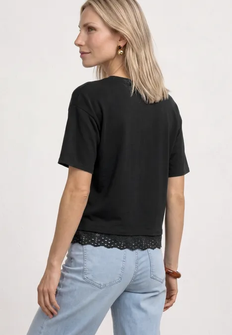 Shirt met ronde hals en kant Black Vrouw van achteren met halflang blond haar, zwart t-shirt met kantzoom en lichtblauwe jeans