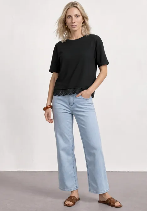Shirt met ronde hals en kant Black Vrouw in zwart T-shirt, lichtblauwe jeans en bruine sandalen voor een neutrale achtergrond.