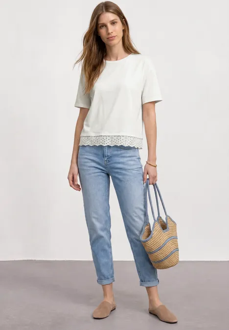 Shirt met ronde hals en kant off white Vrouw met wit kanten topje, blauwe jeans, beige loafers en een strooien tas.