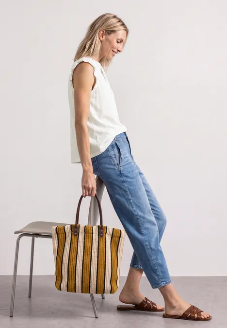 Frau im weißen Top, blauen Jeans und braunen Sandalen, trägt gestreifte Tasche.