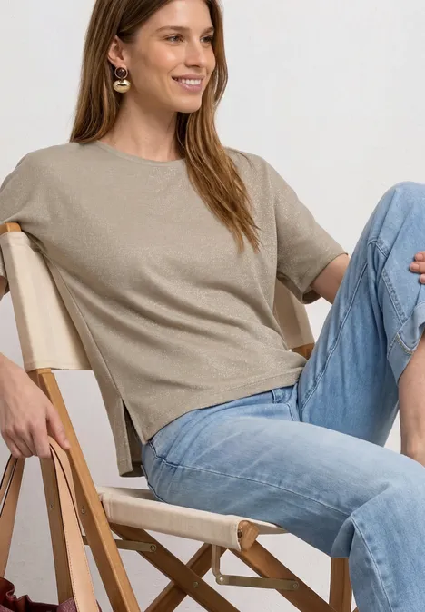 Shirt met overhangende schouders en glitterdetails clay sand Vrouw zit ontspannen op een klapstoel, draagt een beige shirt en blauwe jeans.