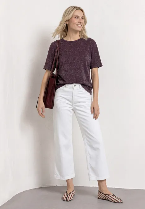 Chemise dropped shoulder avec détails scintillants rustic rouge Une personne porte un t-shirt violet foncé, un jean large blanc, des tongs et un sac bandoulière devant un mur clair.