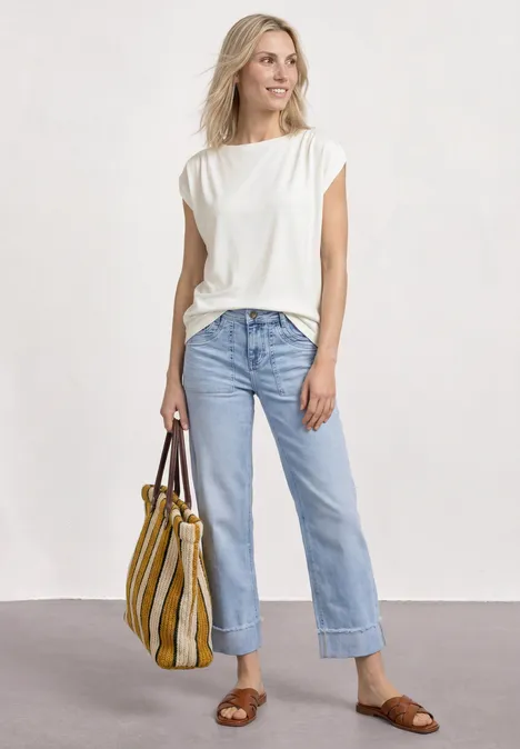 Shirt met overhangende schouders en rimpels off white Vrouw in wit T-shirt, rechte blauwe jeans en bruine sandalen houdt een geel-wit gestreepte tas vast.