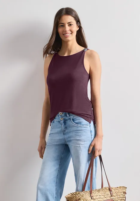 Frau trägt ärmelloses lila Top, helle Jeans und hält eine Strohtasche.
