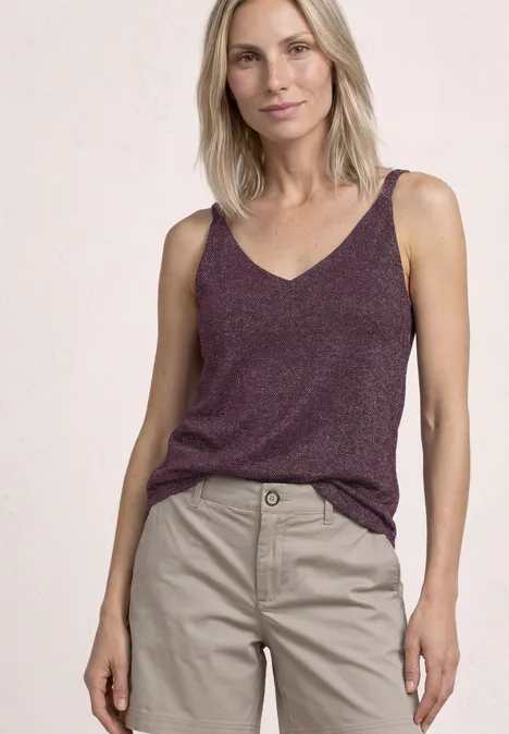 Vrouw met een paarse V-hals top en beige katoenen broek, staand voor een lichte achtergrond.