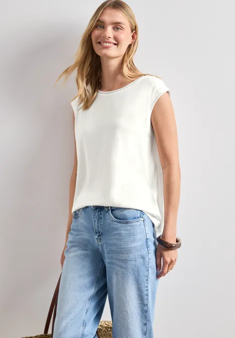 Shirt mit Rundhals und Rippdetails off white Schlanke Frau mit weißem ärmellosem Top und blauen Jeans, hält eine Tasche, vor hellem Hintergrund.