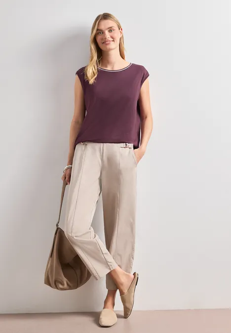 Shirt mit Rundhals und Rippdetails rustic rouge Frau in violettem ärmellosem Oberteil, hellen, weiten Hosen und beigen Schuhen mit passender Tasche.