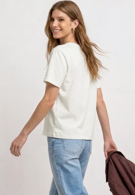 Frau von hinten in weißem T-Shirt, Jeans, hält eine Jacke in der Hand.