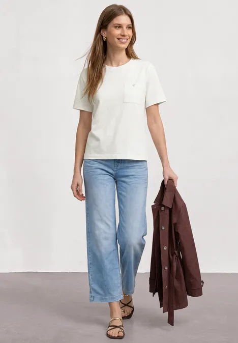 Person trägt weißes T-Shirt, hellblaue, weite Jeans, Sandalen und hält eine braune Jacke.
