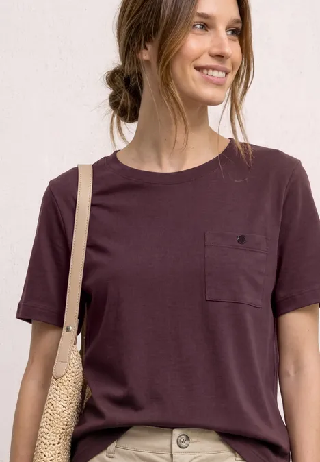 Frau im burgunderroten T-Shirt mit Brusttasche und geflochtenem Schulterbeutel.