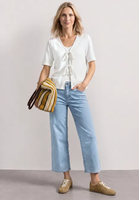 Veste en maille avec rubans off white Femme en top blanc lacé, jean bleu clair, baskets dorées et sac rayé, posant devant un fond neutre.