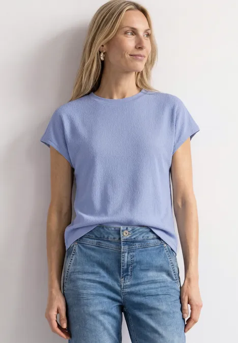 Dropped Shoulder Shirt mit Strukturmix cashmere blue Frau trägt hellblaues T-Shirt und Jeans, steht vor weißem Hintergrund.