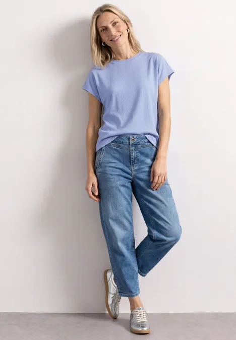 Dropped Shoulder Shirt mit Strukturmix cashmere blue Frau in lässigem Outfit mit hellblauem T-Shirt, lockeren Jeans und Turnschuhen lehnt entspannt an Wand.