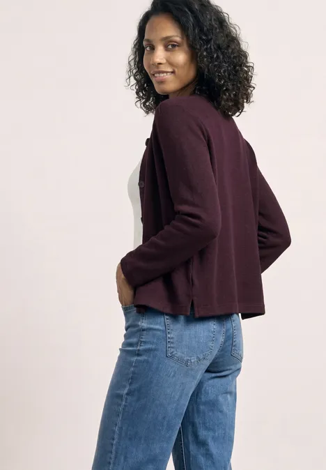 3/4-Arm Jacke in Strick-Optik rustic rouge Frau mit lockigem Haar trägt eine burgunderfarbene Strickjacke und blaue Jeans, seitlich stehend.