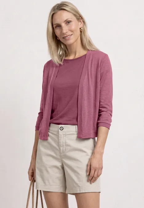 Blonde Frau in rosa Shirt und Cardigan mit beigen Shorts und passender Tasche vor weißem Hintergrund.