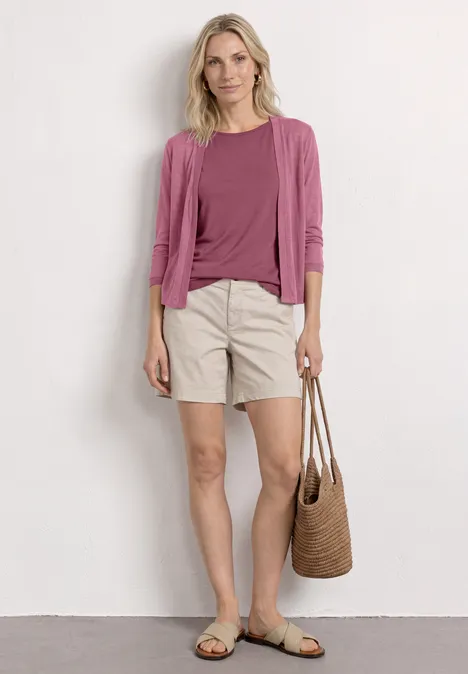 Frau in rosa Oberteil und Cardigan, beige Shorts, Sandalen und Strohtasche, lässiger Sommerlook.