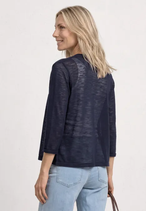Vrouw van achteren in een marineblauw licht vest met driekwarts mouwen en een lichte spijkerbroek, met tas in de hand.