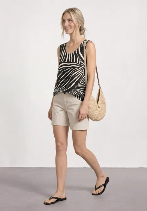 Top met V-hals en elastische zoom Black Vrouw in zebratop, beige short, strooien tas en slippers, lopend voor een witte muur.