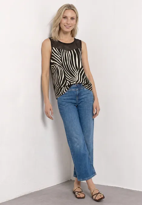 Tup mit Rundhals und Mesh-Einsatz Black Eine junge blondhaarige Frau lehnt vor einer weißen Wand, trägt ein Zebra-Top, gerade Jeans und Sandalen.