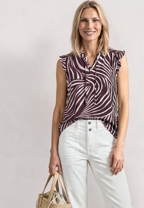 Een vrouw draagt een mouwloze blouse met zebraprint, een witte broek en houdt een strooien tas vast.