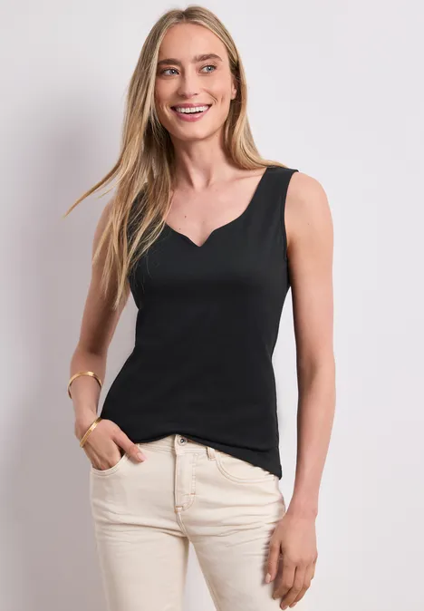 Basic Top mit Herzausschnitt Black Frau trägt ärmelloses schwarzes Oberteil und helle Hose, steht seitlich vor neutralem Hintergrund.