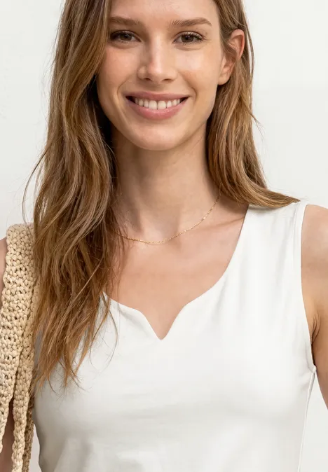 Basic Top mit Herzausschnitt off white Frau in weißem ärmellosem Oberteil mit V-Ausschnitt, zarter Halskette und gehäkelter Tasche.