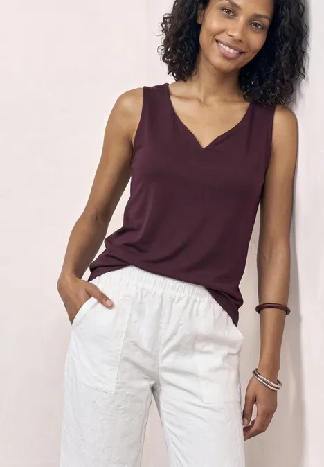 Une personne porte un haut sans manches bordeaux et un pantalon blanc sur fond clair.