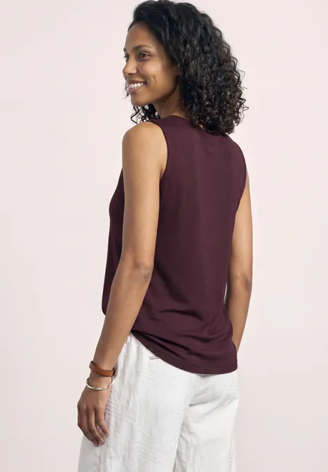 Personne de dos, visage flou, cheveux bouclés, porte un top bordeaux sans manches et un pantalon blanc.