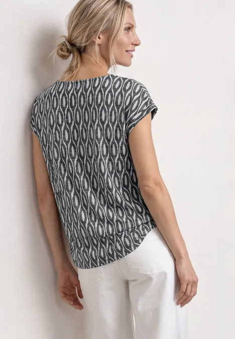 Femme de dos, porte un T-shirt noir et blanc à motif graphique et un pantalon blanc, appuyée contre un mur.