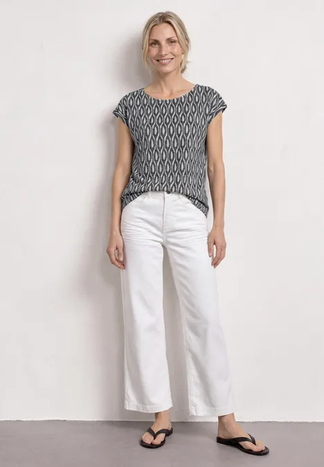 Jeune femme en haut à motif, pantalon blanc ample et tongs noires devant un mur blanc.