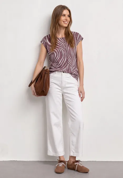 Femme en pantalon blanc ample, top rayé violet et sac marron, debout devant un mur blanc.