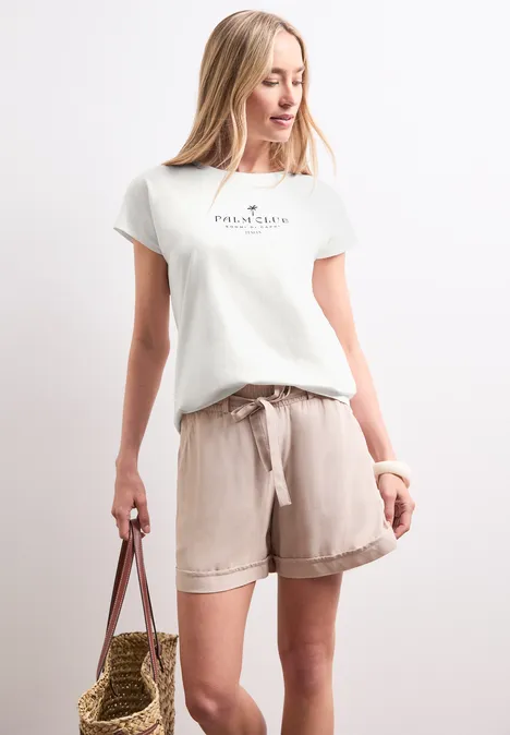 Shirt met hangende schouder en tekst off white Vrouw in wit t-shirt met opschrift, beige linnen short en strooien tas voor een witte achtergrond.