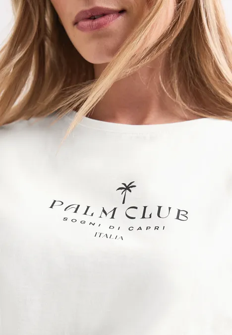 Shirt met hangende schouder en tekst off white Close-up van een wit t-shirt met zwarte tekst ‘Palm Club Sorrento, Capri Italia’ en een palmboomlogo.